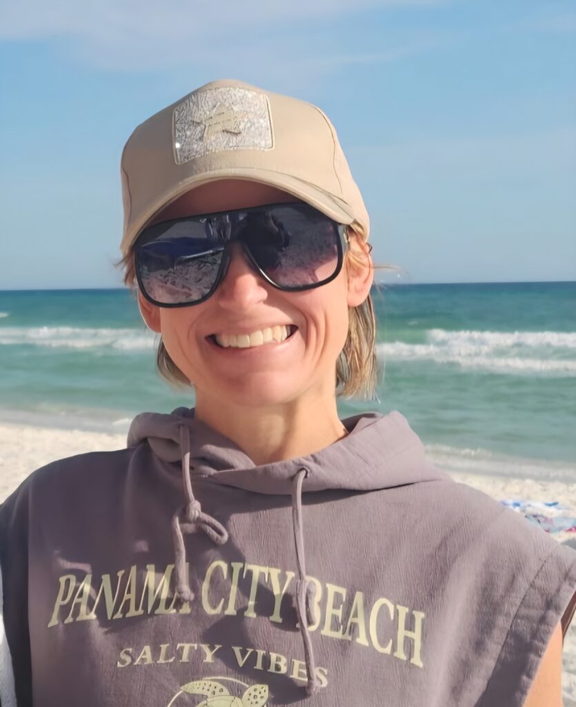 Allyson Bennett, smiling, with a tan hat, black sunglasses, and a PCB shirt at the beach